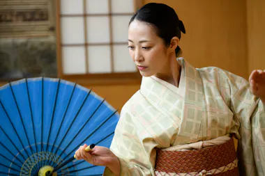 Geisha in a pale kimono dancing with a blue traditional Japanese umbrella at a private show in Shibuya, Tokyo