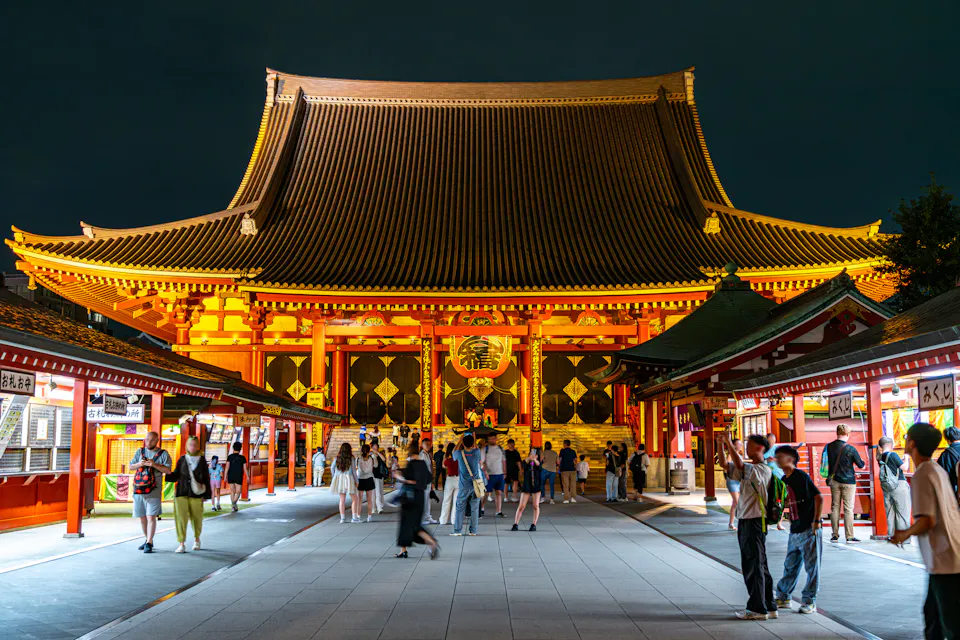 Senso-ji Temple's main hall lit at night in Asakusa, with visitors walking along the stone approach between shop buildings