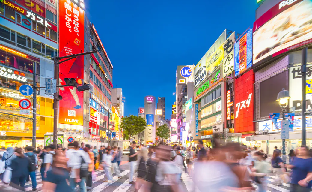 Crowds cross the scramble intersection in Shibuya at dusk, surrounded by lit billboards and shopping buildings