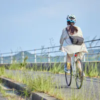 A rider on a riverside cycling path, the easiest kind of riding in Tokyo. A cyclist in a white helmet and light jacket rides away from the camera along a paved riverside path lined with tall grass