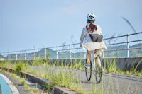 A cyclist in a white helmet and light jacket rides away from the camera along a paved riverside path lined with tall grass