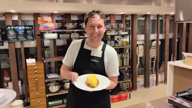 Guest holding a plate of omurice in a cooking studio lined with shelves of cookware