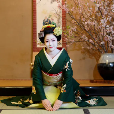 A maiko in a green kimono greets guests with a bow. Maiko in a green kimono kneeling and bowing beside a vase of cherry blossoms