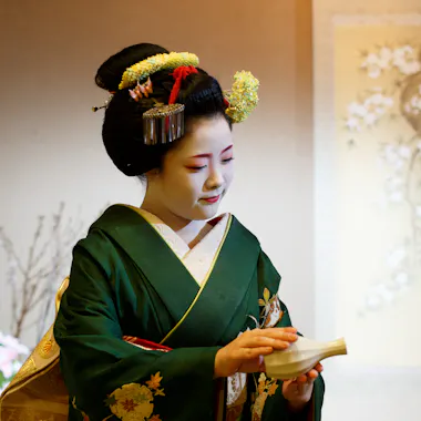 A maiko with a ceramic sake flask. Maiko in a green kimono and floral hair ornaments holding a ceramic sake flask