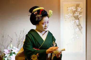 Maiko in a green kimono and floral hair ornaments holding a ceramic sake flask