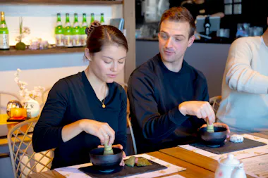 Couple whisking matcha in black tea bowls at a wooden table
