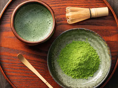 Bowl of whisked matcha beside matcha powder and a bamboo whisk