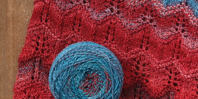 A blue and red ball of yarn sits on top of a red and blue lace cowl.