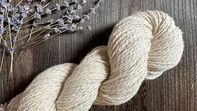 A skein of beige-colored alpaca yarn with dried stems of lavender on a wood background.