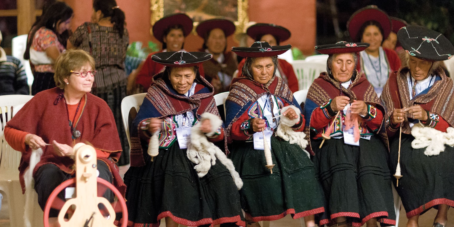 Seated woman at spinning wheel and 4 spinners in traditional costume spinning on low-whorl spindles.