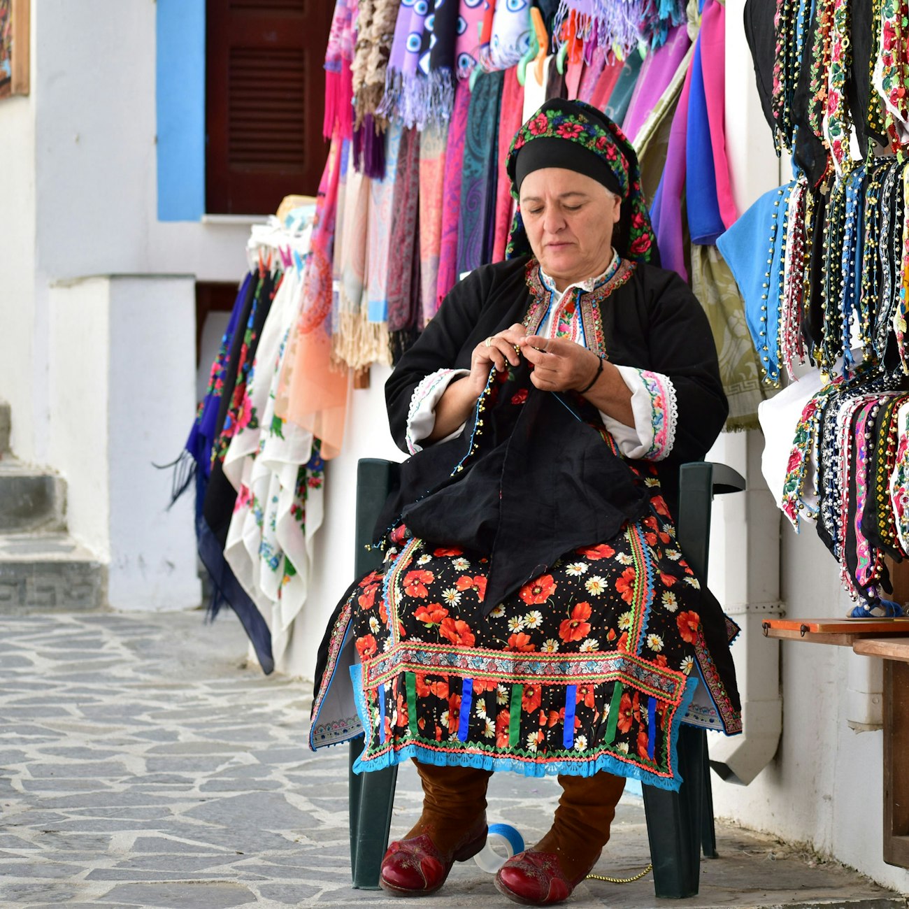Woman in Greek costume (black headscarf, black cardigan trimmed with ribbon, colorful skirt) sits on a walkway and stitches an edging on a handkerchief.