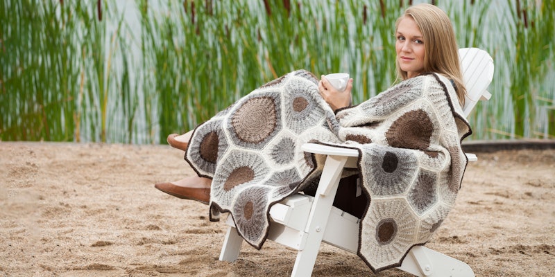 Woman with light brown hair sitting in an adirondack chair on a sandy beach with brown, gray, and cream afghan draped across her