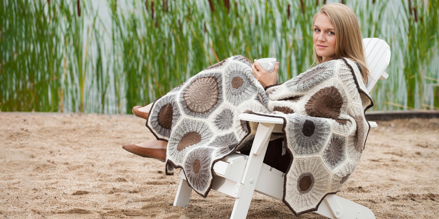 Woman with light brown hair sitting in an adirondack chair on a sandy beach with brown, gray, and cream afghan draped across her