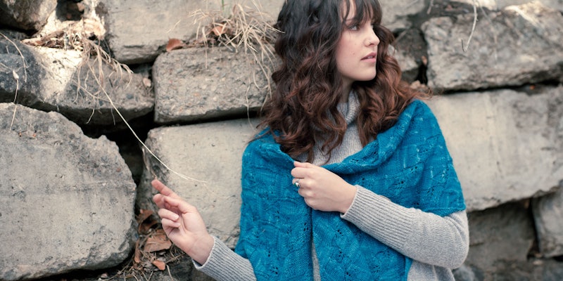 Woman with dark hair wearing teal entrelac shawl standing agains stone wall