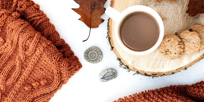 Brown cable knitting and gray stones with brown beverage in mug and cookies on tree stump