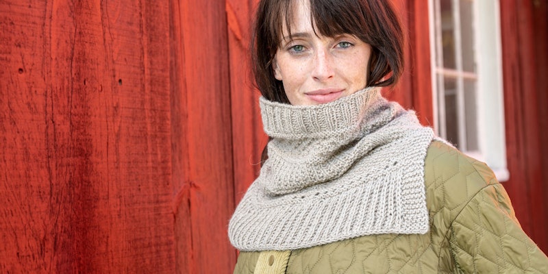 woman with brown hair wearing sand-colored knitted cowl standing against a red barn