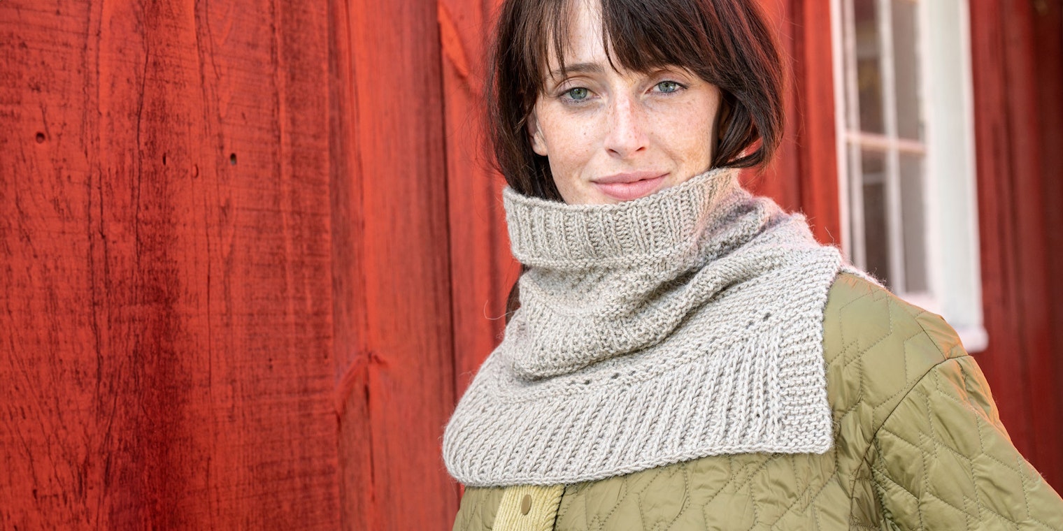 woman with brown hair wearing sand-colored knitted cowl standing against a red barn