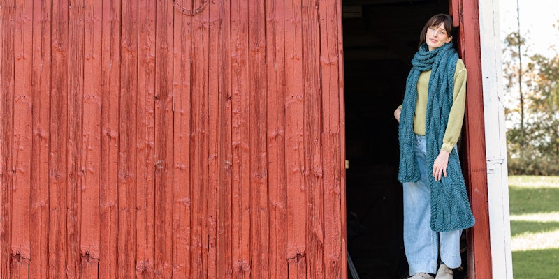 Model with large teal knitted scarf standing in doorway of red barn