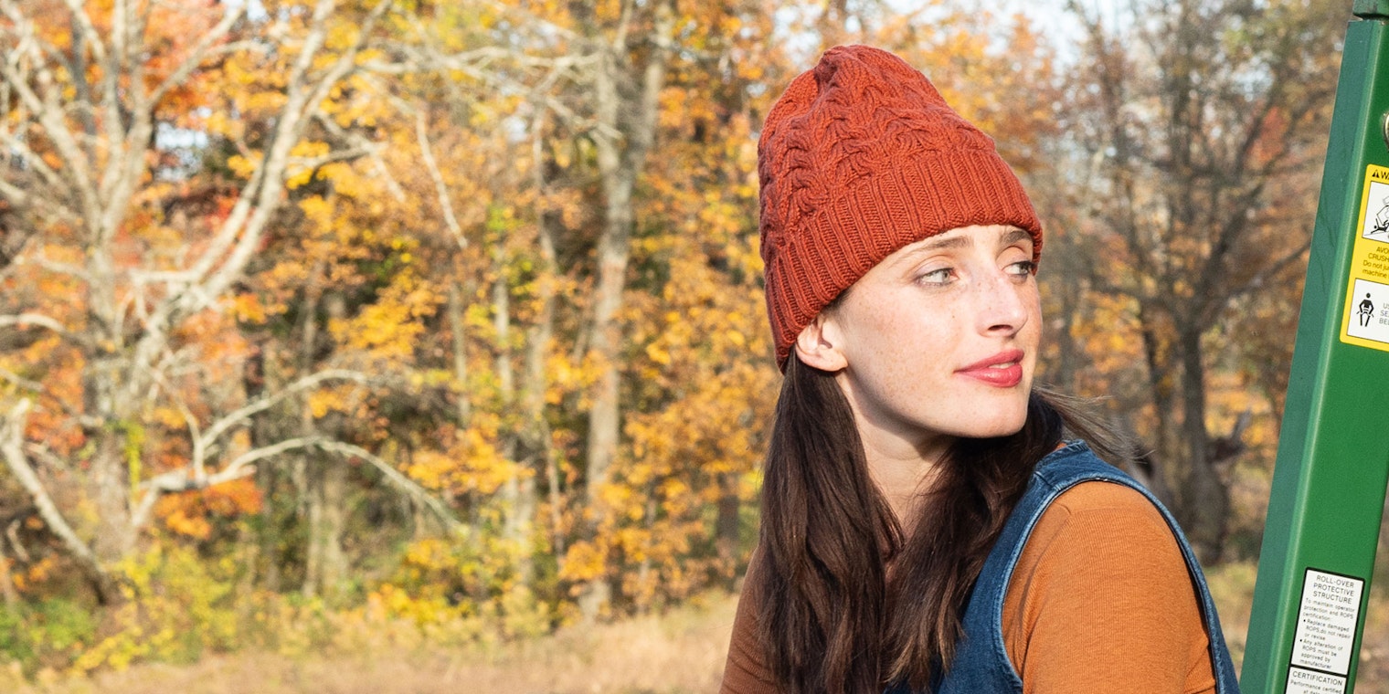 Partial profile of model seated on tractor wearing orange cabled hat