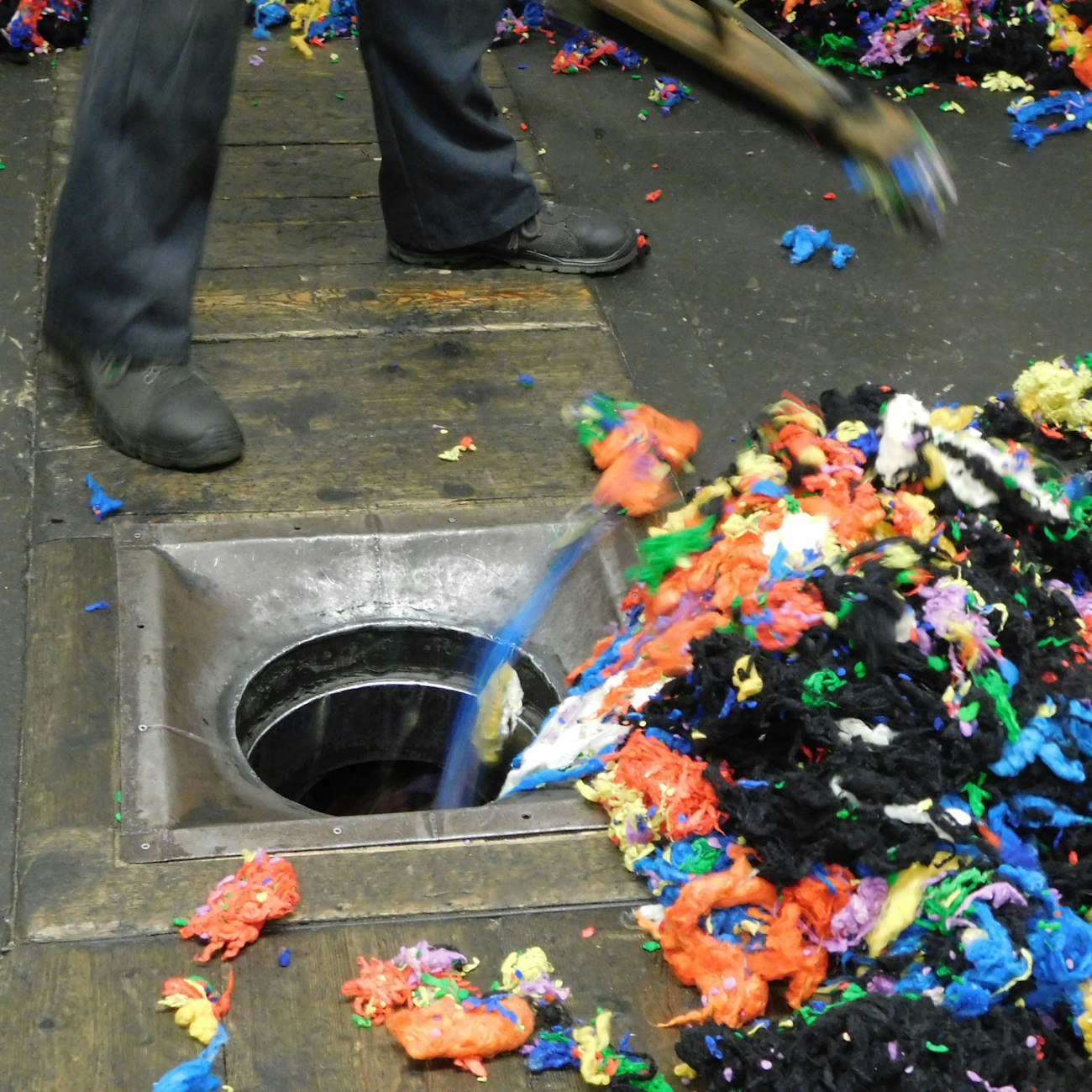 Person in dark shoes moves a pile of multicolored wool scraps into an iron-ringed circle on the floor