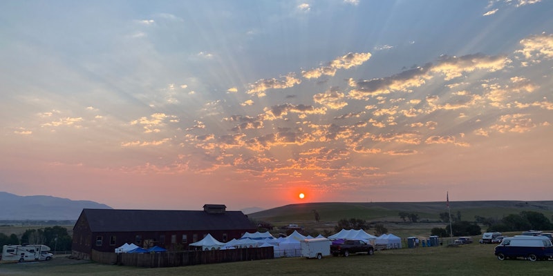 Long brown barn and white tents in field, campers around, as the sun rises