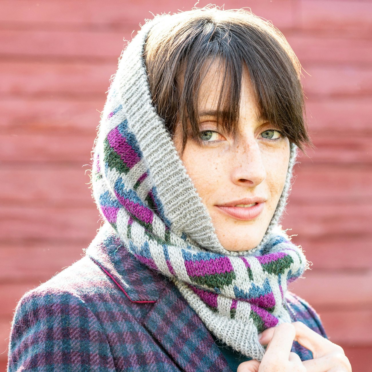 Woman in gray knitted cowl with blue, pink, and green flowers, standing in front of a red barn