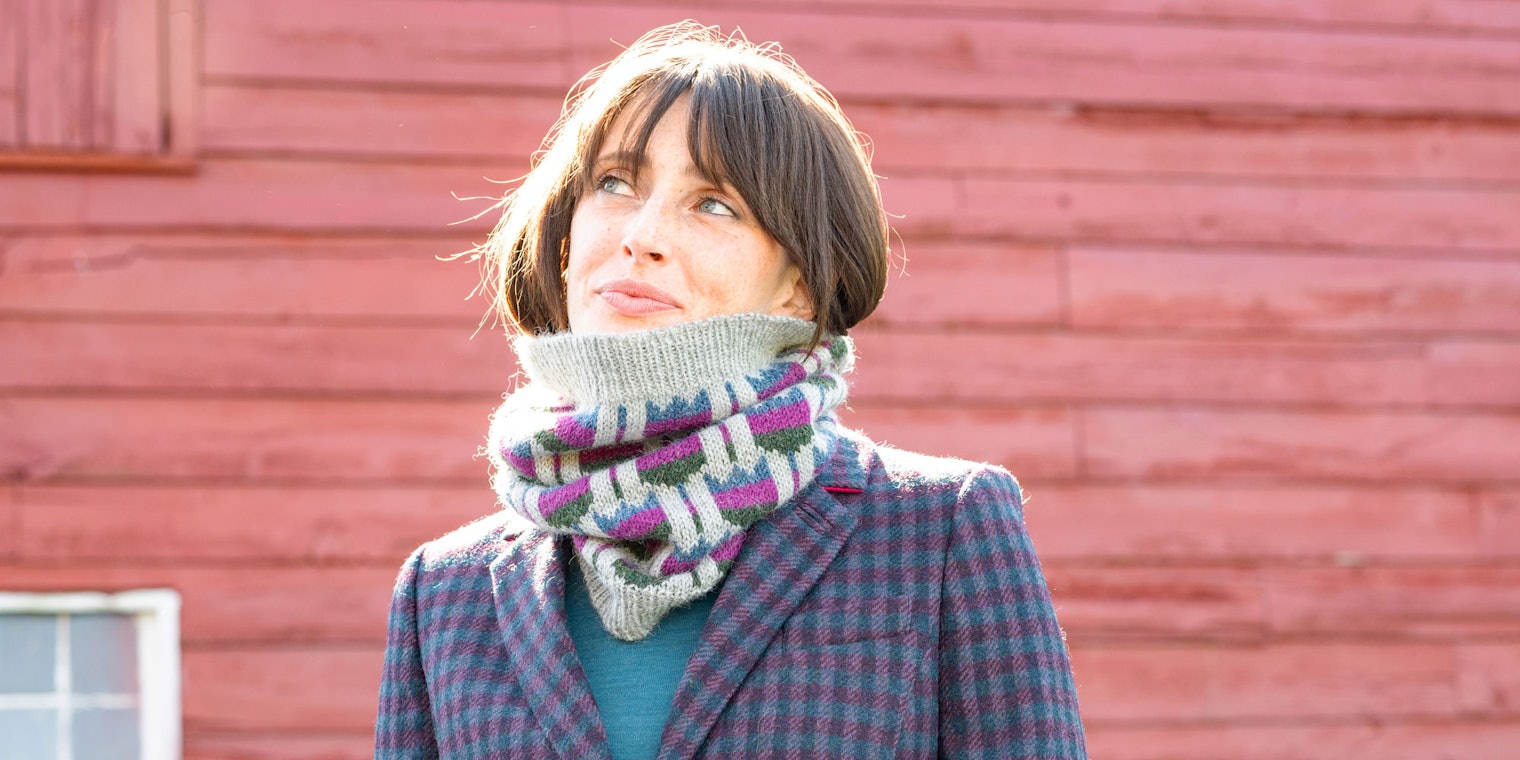 Woman in gray knitted cowl with blue, pink, and green flowers, standing in front of a red barn