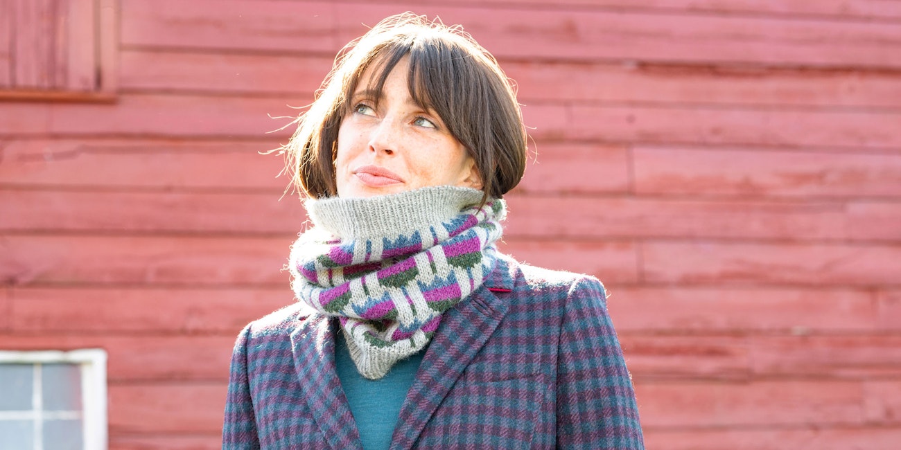 Woman in gray knitted cowl with blue, pink, and green flowers, standing in front of a red barn