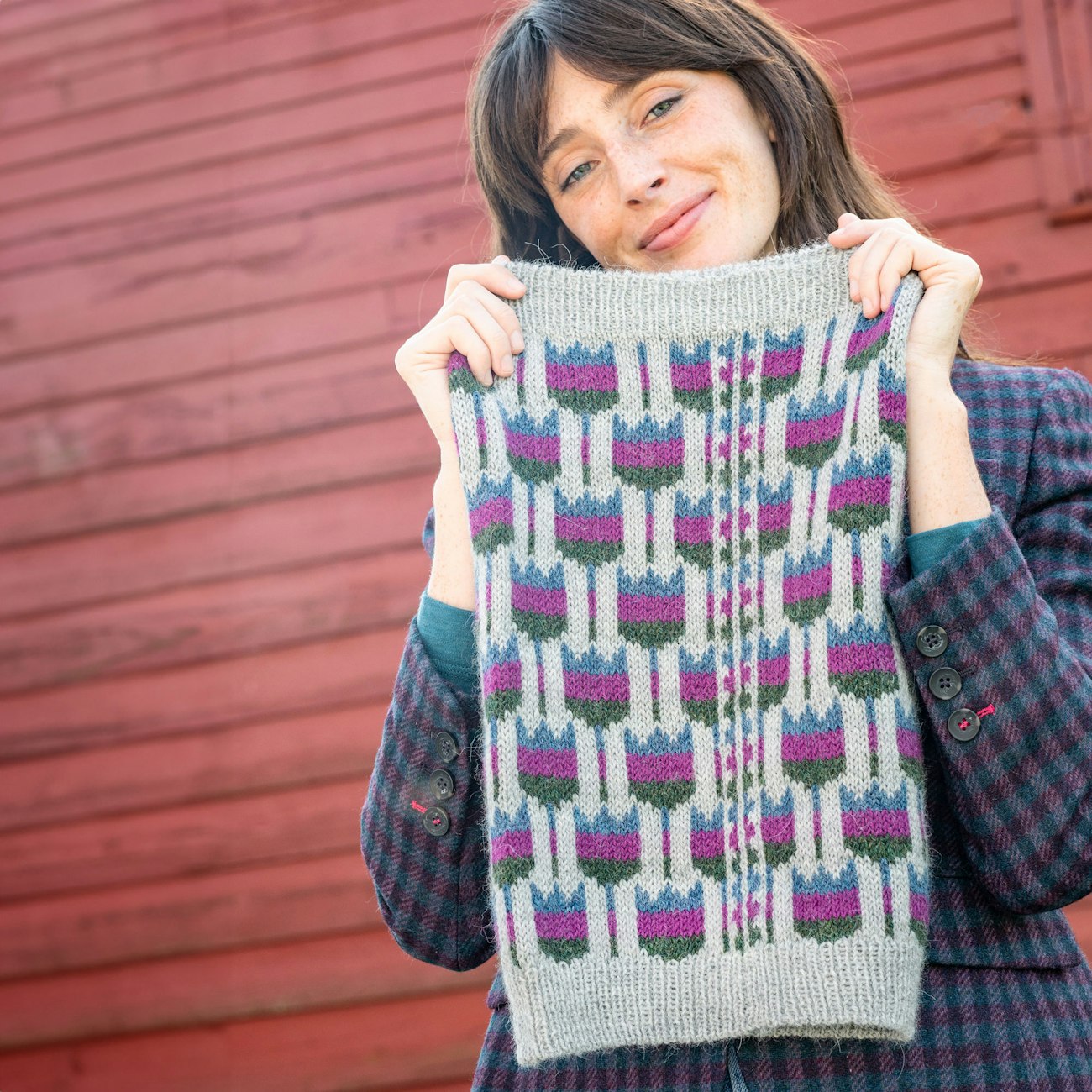 Woman in gray knitted cowl with blue, pink, and green flowers, standing in front of a red barn