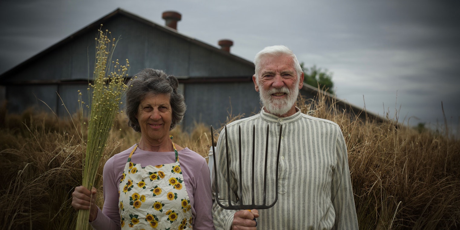 Woman holding flax and man holding pitchfork stand in front of barn in style of American Gothic painting