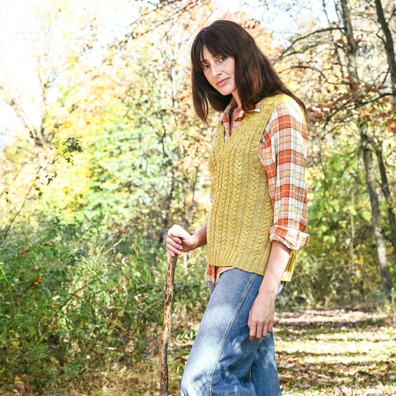 Woman with long brown hair, gold cabled knitted vest, plaid shirt, and jeans carries walking stick in woods