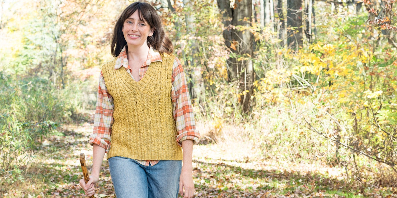 Woman with long brown hair, gold cabled knitted vest, plaid shirt, and jeans carries walking stick in woods