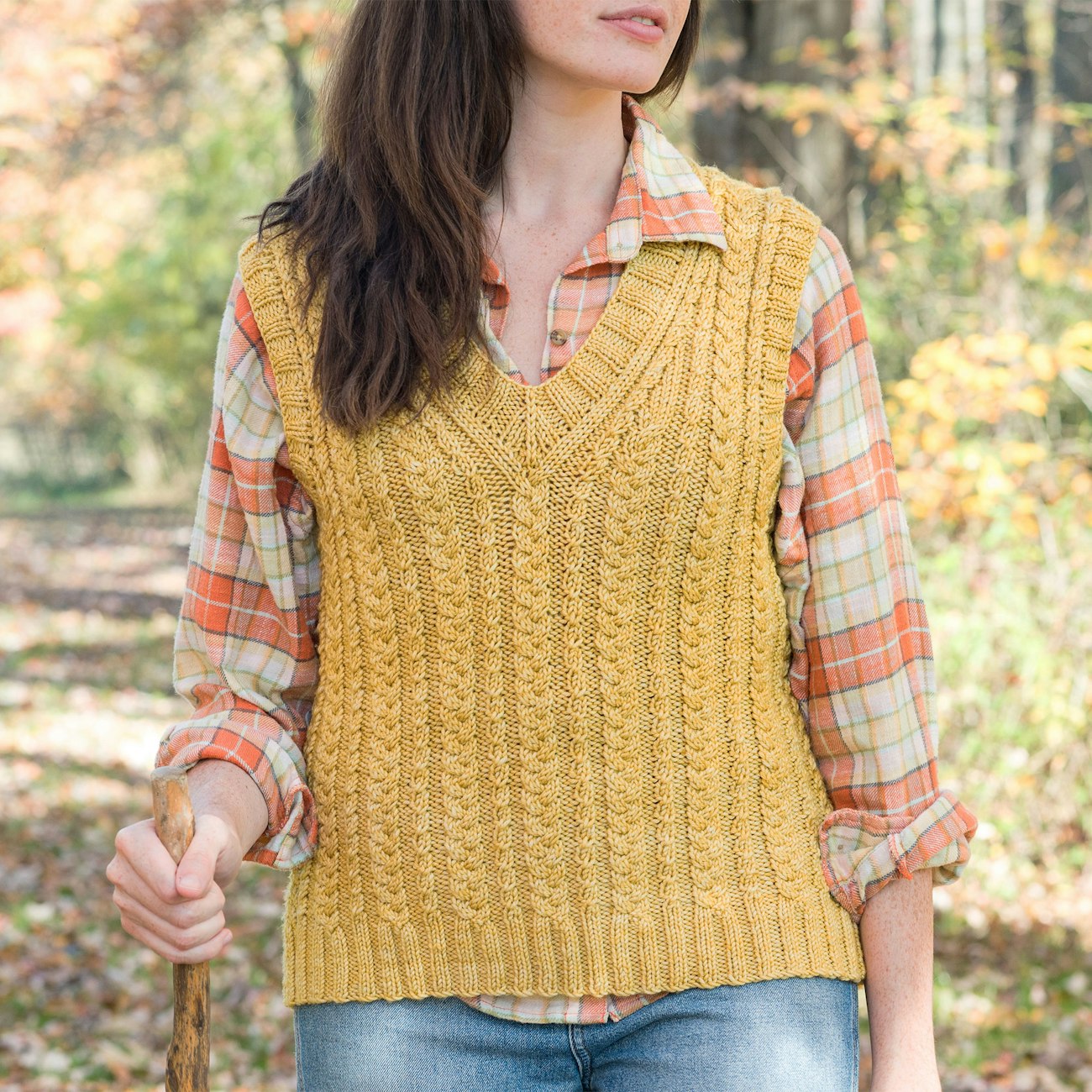 Woman with long brown hair, gold cabled knitted vest, plaid shirt, and jeans carries walking stick in woods