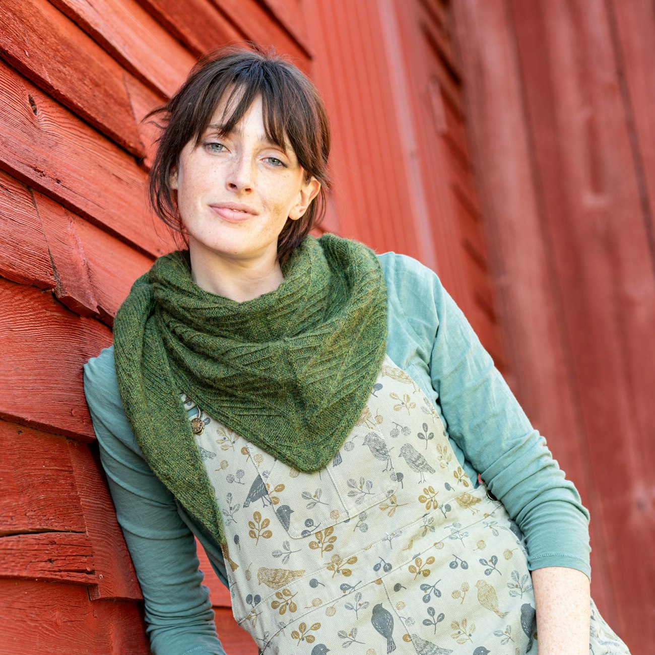 Woman with brown hair wearing dark green knitted shawl standing against red barn