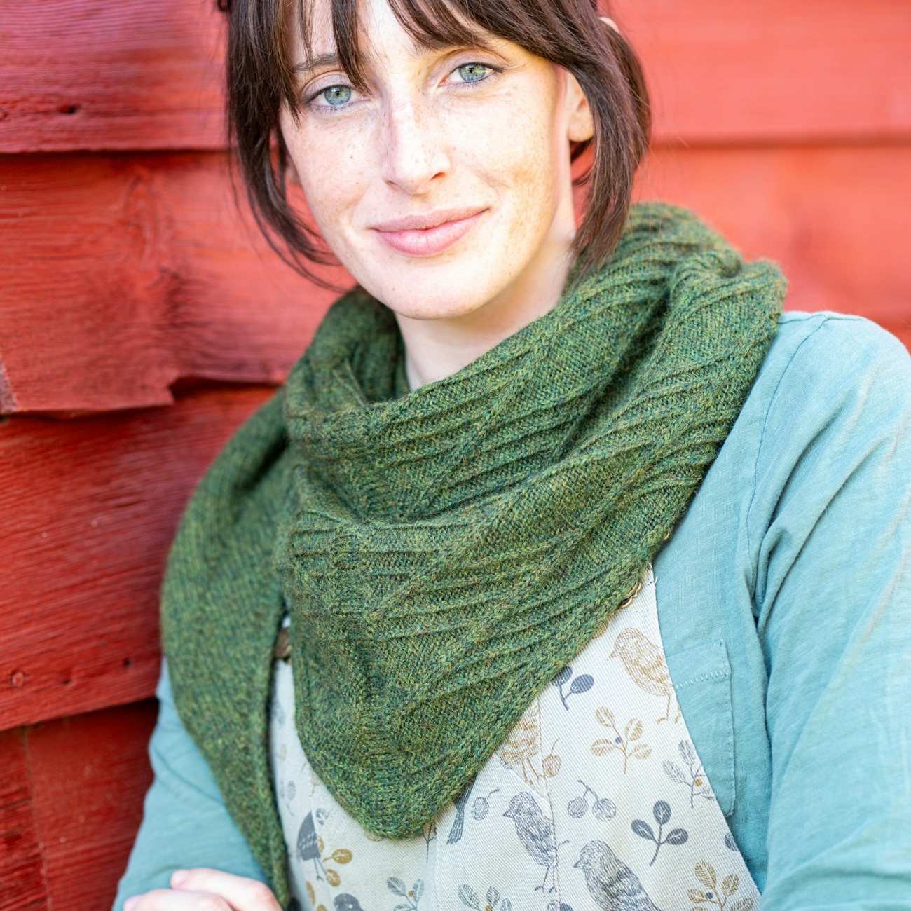 Woman with brown hair wearing dark green knitted shawl standing against red barn