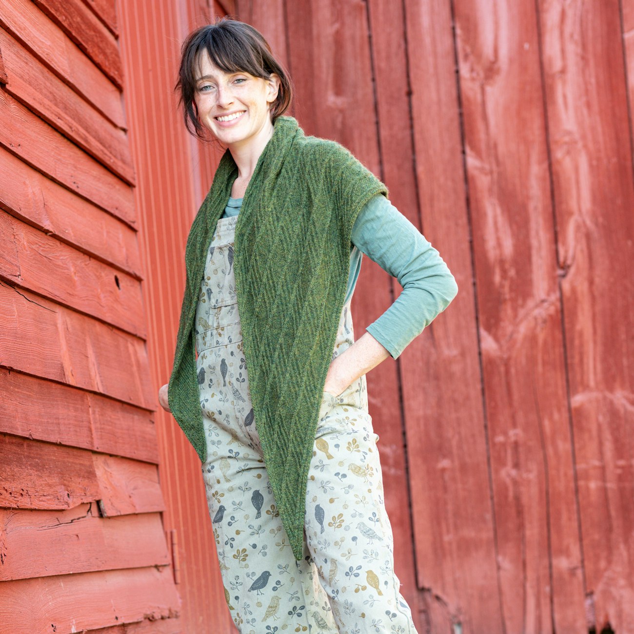 Woman with brown hair wearing dark green knitted shawl standing against red barn
