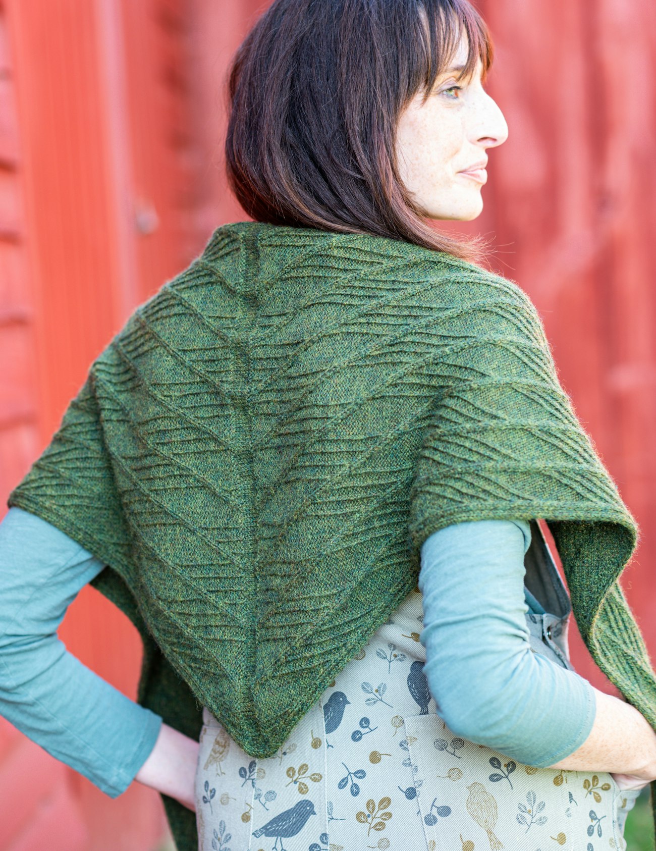 Woman with brown hair wearing dark green knitted shawl standing against red barn