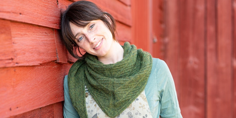 Woman with brown hair wearing dark green knitted shawl standing against red barn