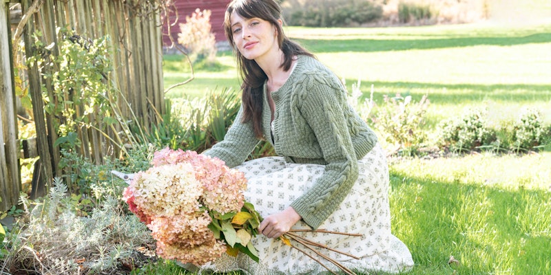 Woman in sage-green cardigan and flowered dress, crouching on the ground and holding dried flowers
