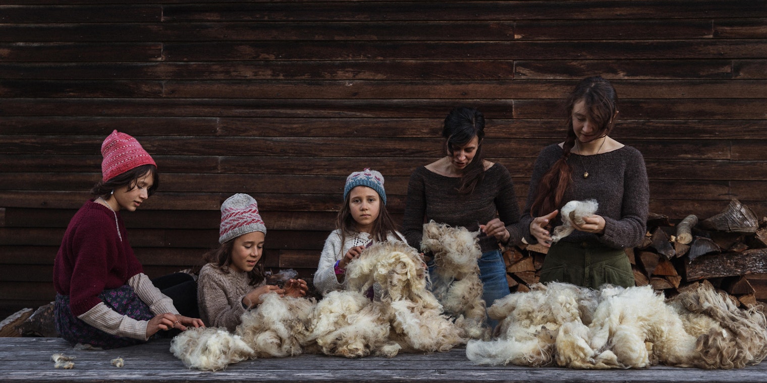 Three children in handknitted wool hats, one teenager, and one adult woman sorting raw wool