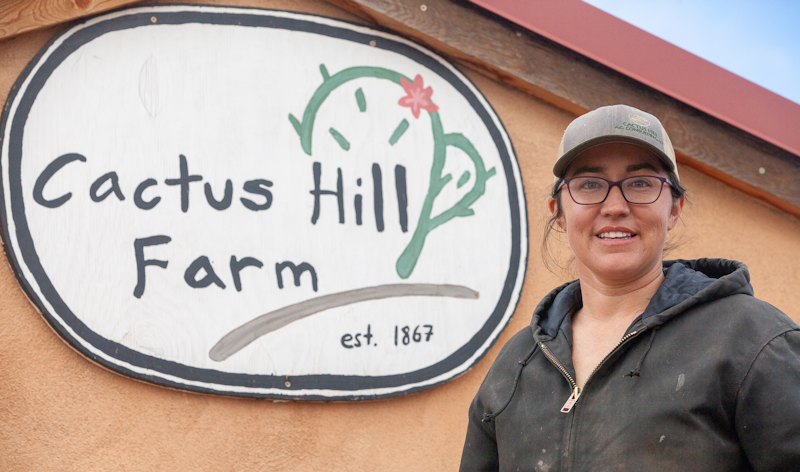 Woman in black jacket, baseball cap, and glasses standing next to a sign that reads Cactus Hill Farm
