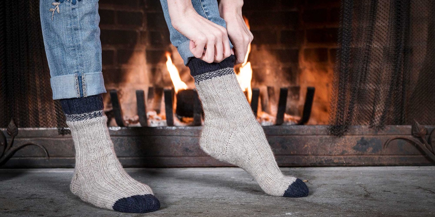 A person standing on a grey stone hearth in front of a fire. The person is shown from the calf-down and is pulling on a pair of light grey socks with navy blue accents at the cuff and toe.