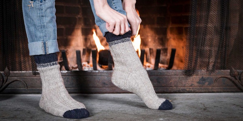 A person standing on a grey stone hearth in front of a fire. The person is shown from the calf-down and is pulling on a pair of light grey socks with navy blue accents at the cuff and toe.