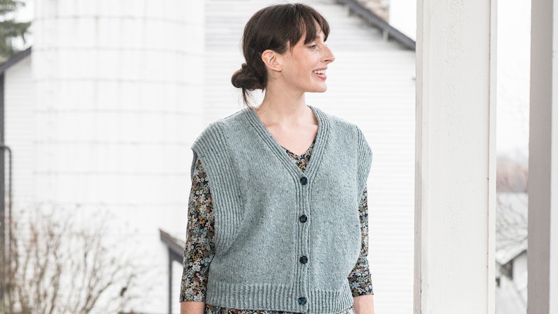 A person standing in front of a white barn, looking off into the distance. The person is wearing a loose blue-gray vest over a flowered dress.