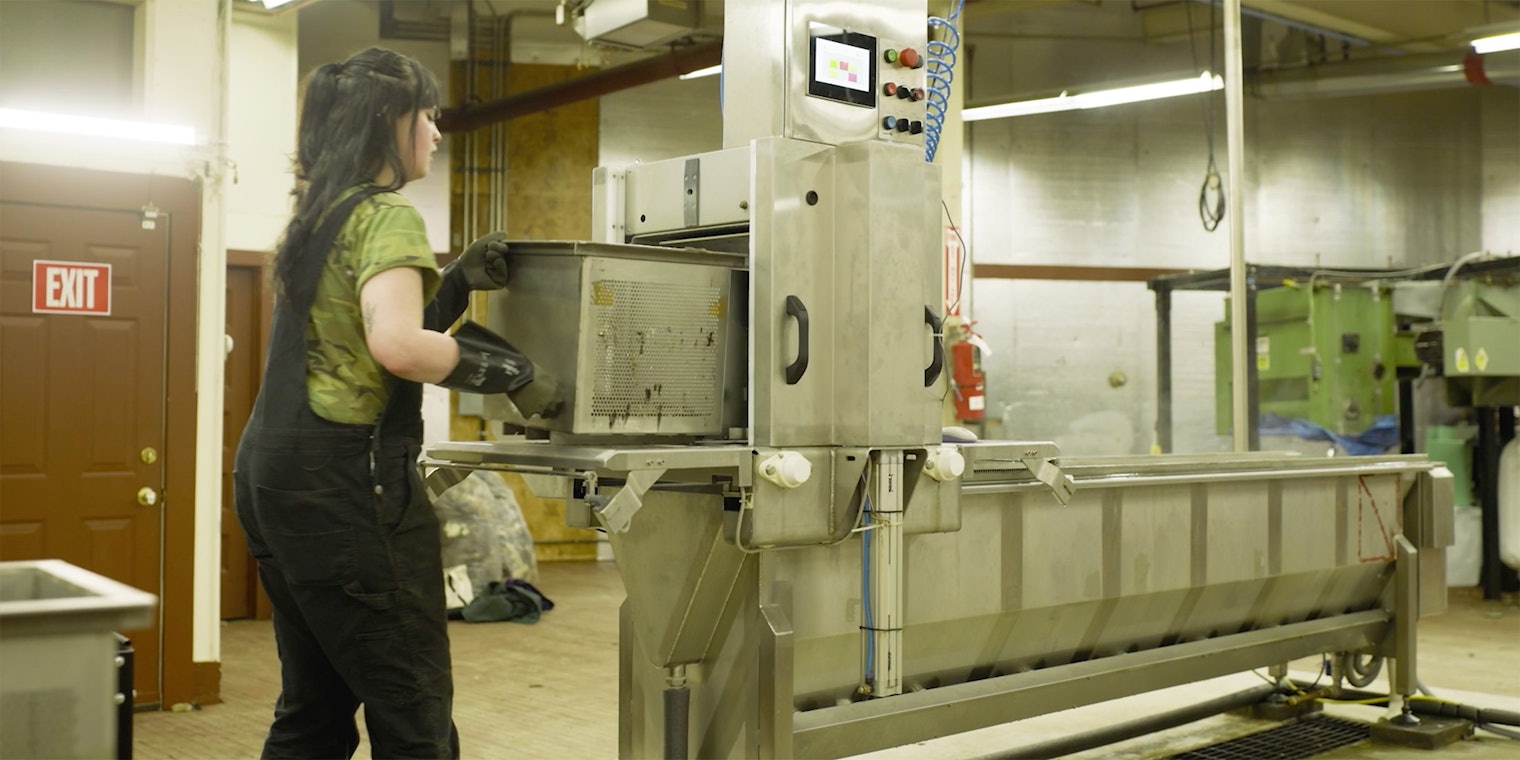 Woman in black overalls removes a metal mesh basket from a steaming silver machine