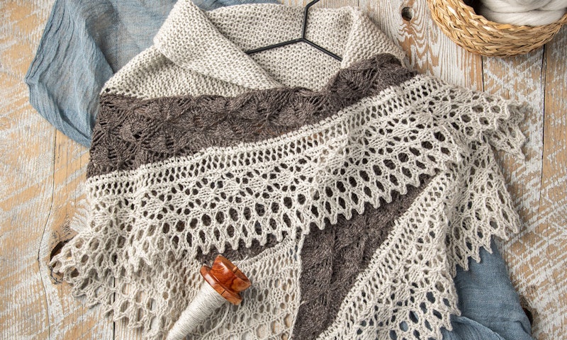 A shawl in different shades of gray rests on a distressed wood table.