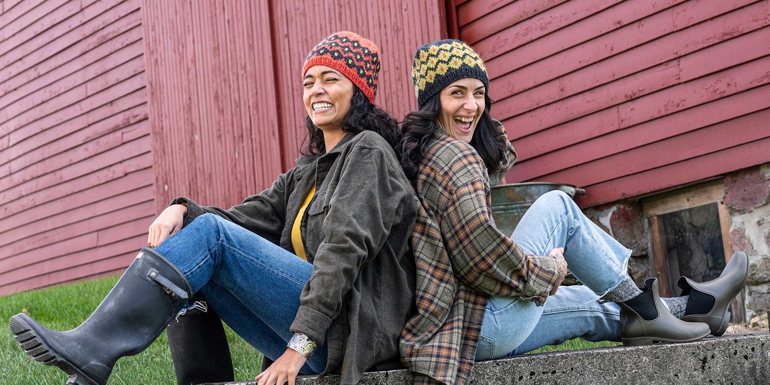 Two women sit back-to-back in front of a red barn. They are wearing hats with diamond motifs and laughing.