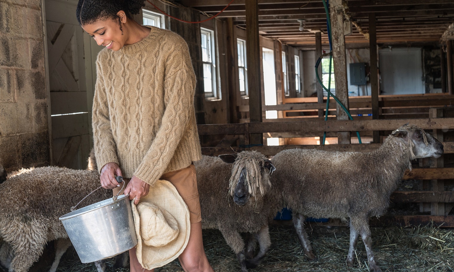 Woman in tan pullover with silver bucket in barn with sheep