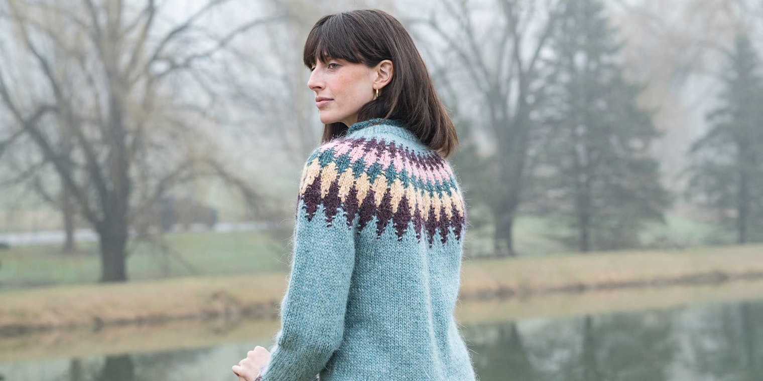 A woman wearing a patterned Icelandic Lopi sweater stands in front of a foggy pond.