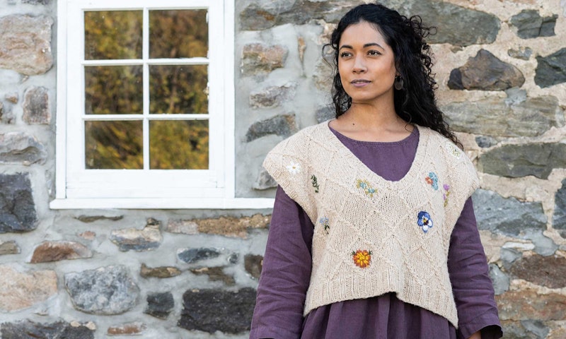 woman standing near a stone wall wearing a purple dress and knitted vest